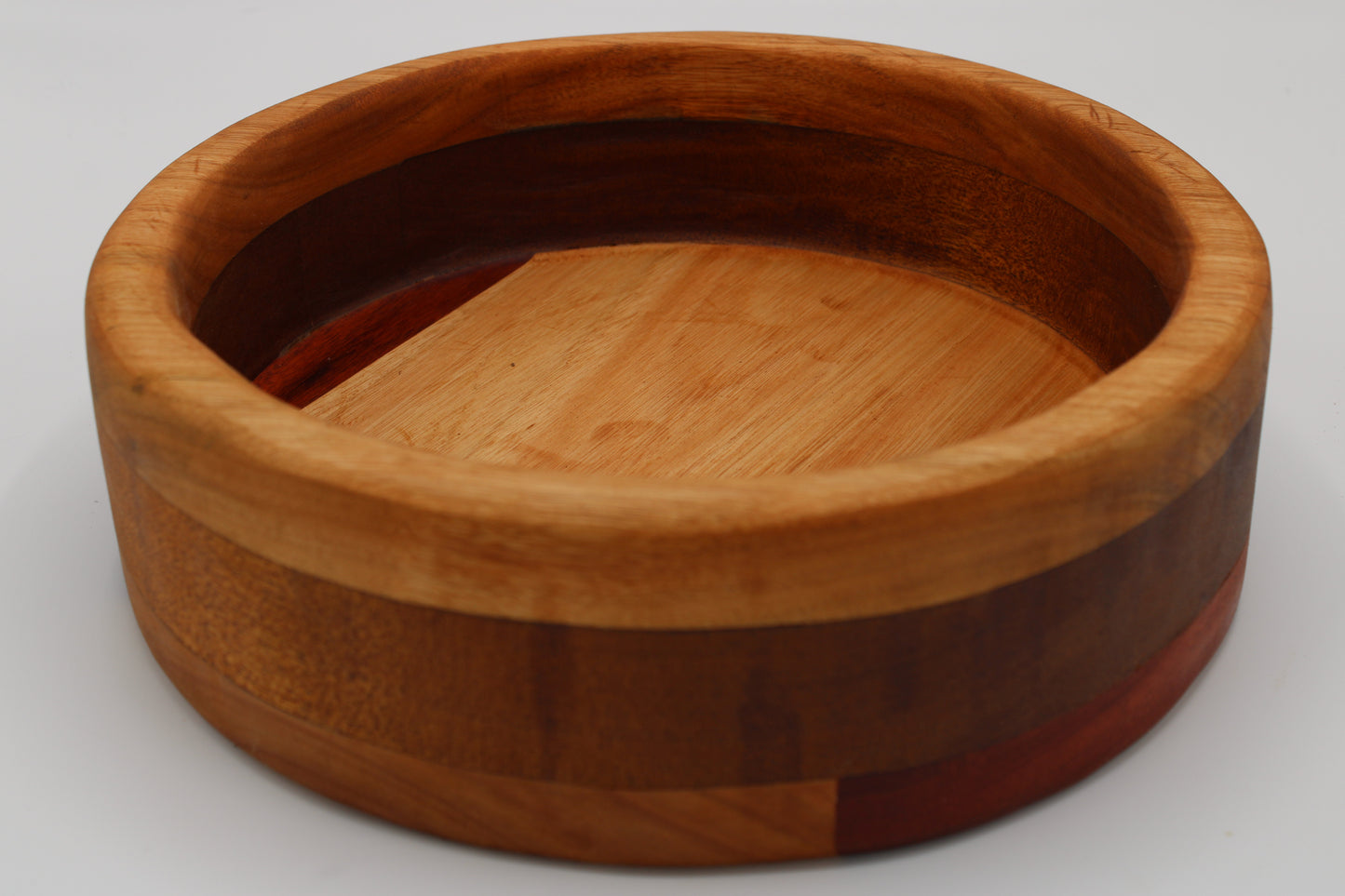 A round wooden bowl with a multi-toned wood grain design, featuring a light tan outer rim, a medium brown center, and dark reddish-brown accents on the sides, photographed on a light gray background.
