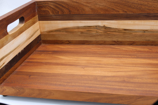 Close-up of a wooden serving tray or board with a cutout handle on the side, showing layered wood grain in warm brown and tan tones.