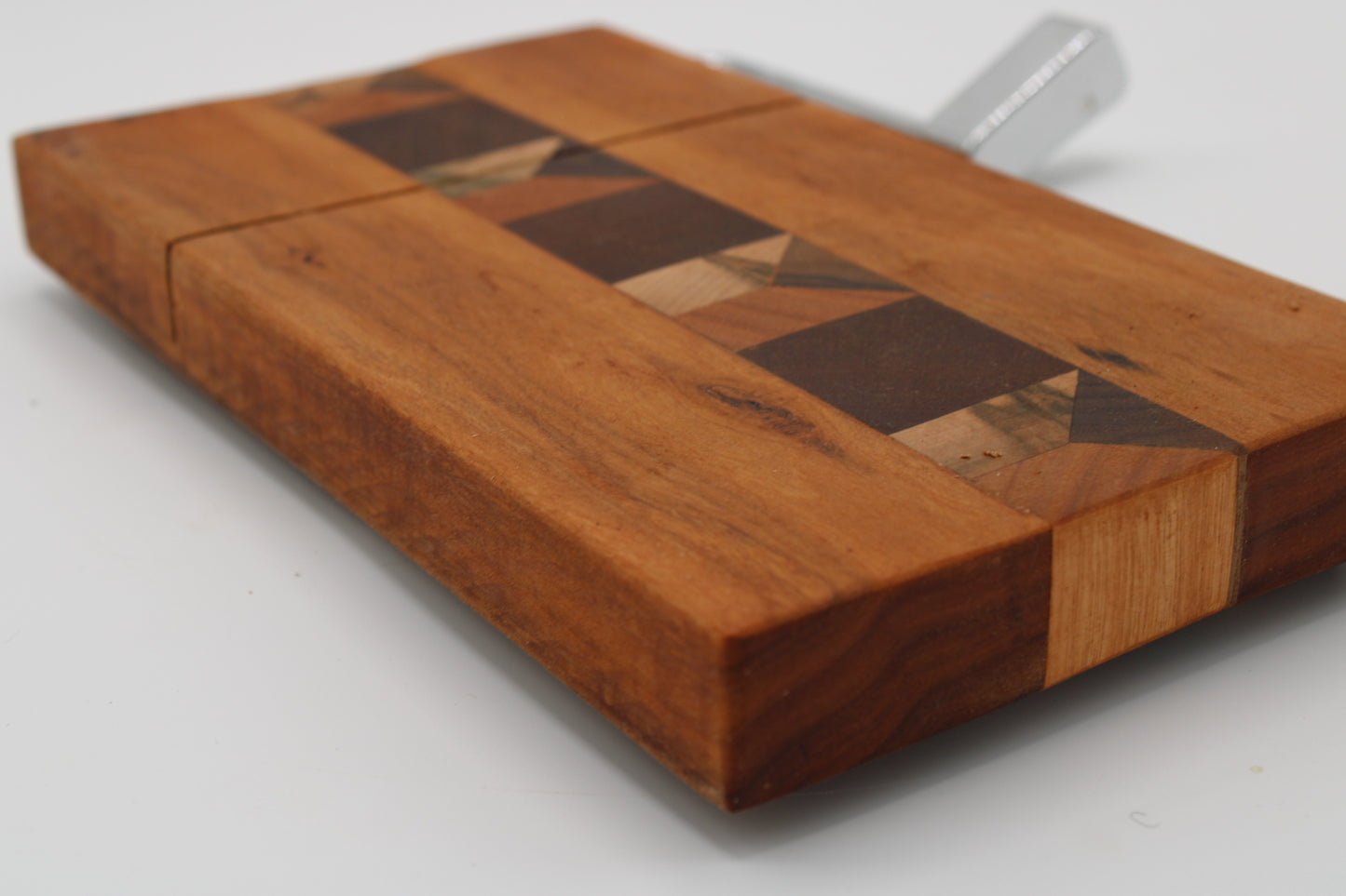 A close-up of a wooden cheese board with a geometric inlay pattern in medium and dark brown wood tones, featuring a silver metal clasp on a white background.