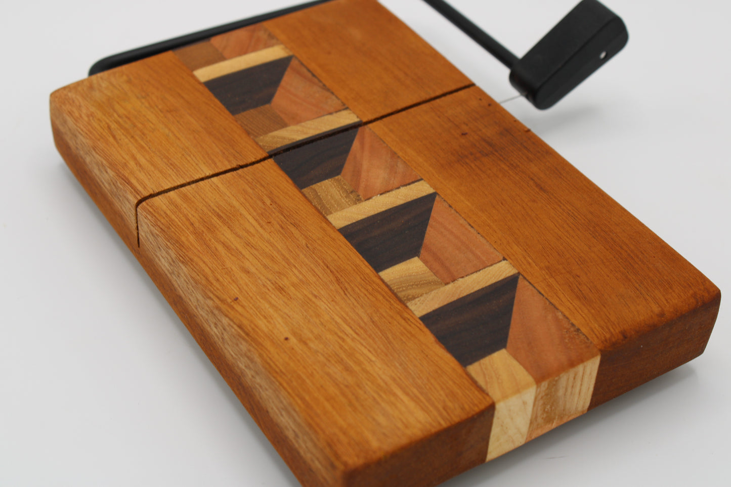 A close-up of a wooden cheese board with a geometric inlay pattern in medium and dark wood tones, shown on a light gray background with a black handle or tool visible at the top right.