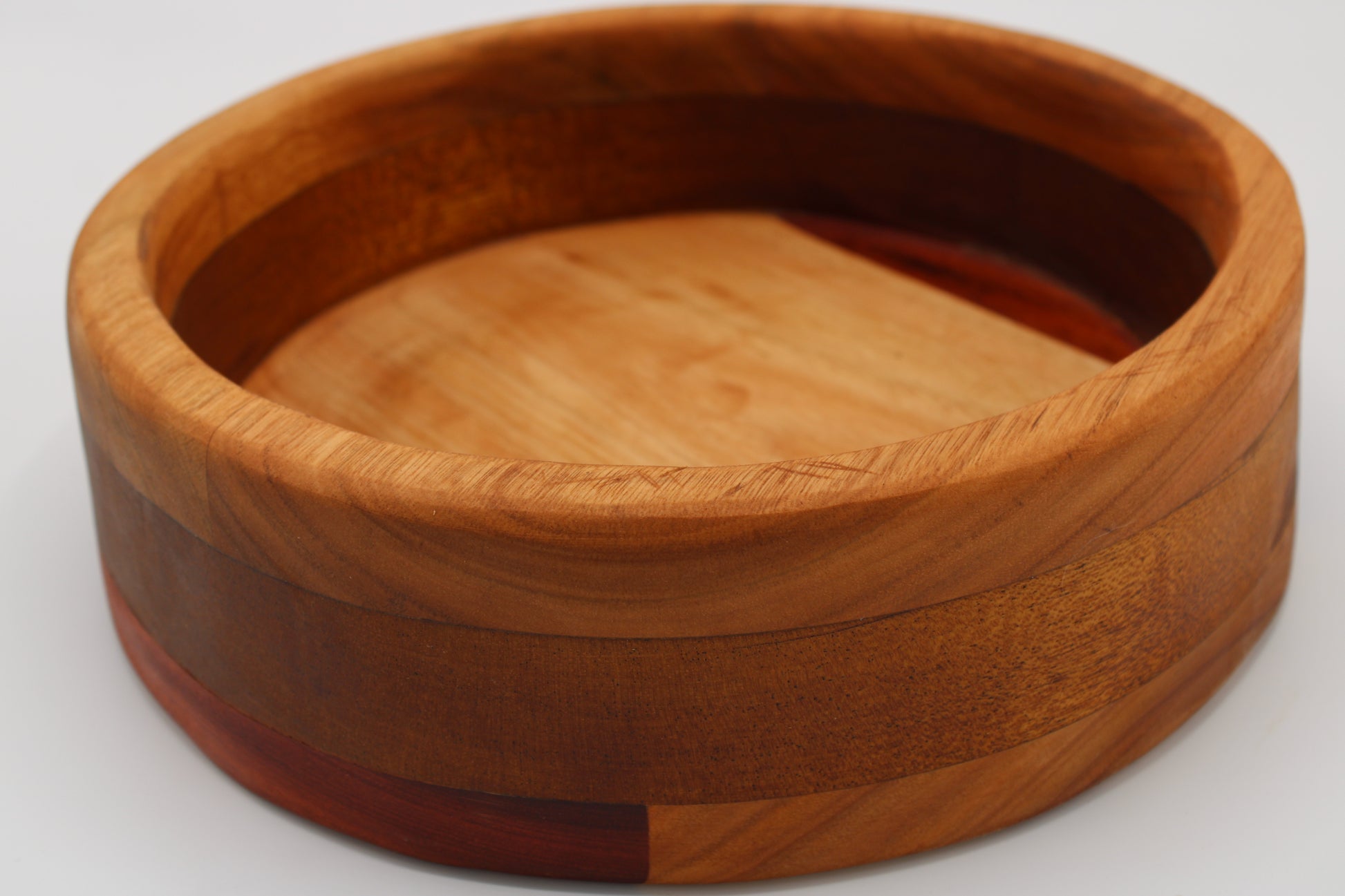 A round wooden bowl with a thick rim, crafted from layered wood strips in warm tan, medium brown, and reddish accents, shown on a light gray background.