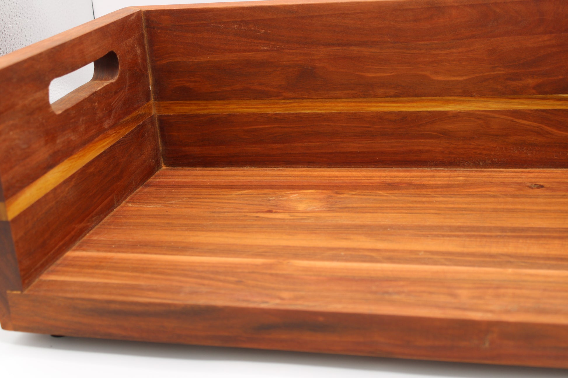 Close-up of a wooden tray or shelf with a warm reddish-brown finish and a cutout handle on the left side, photographed against a light neutral background.