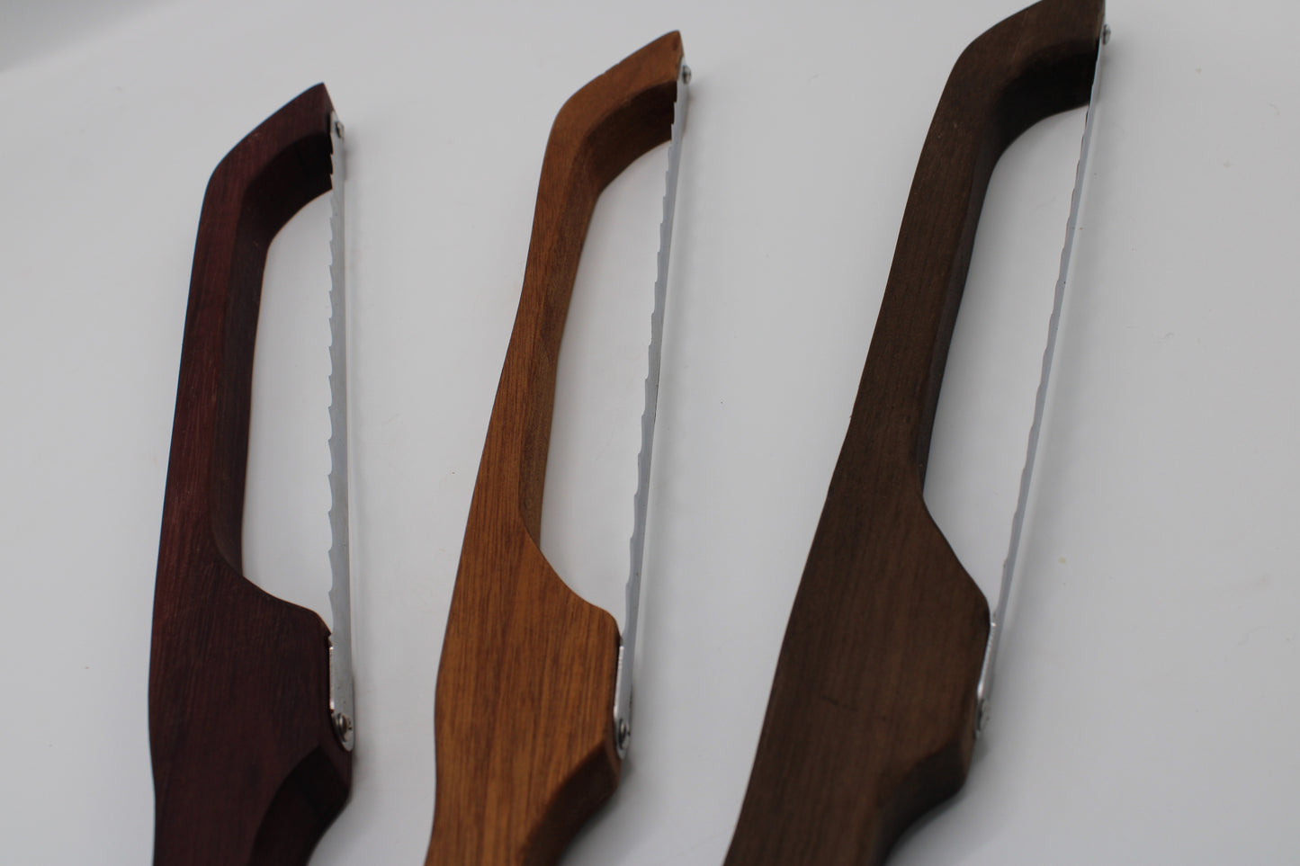 Three wooden-handled folding knives with serrated stainless steel blades, shown in three different wood finishes against a plain white background.