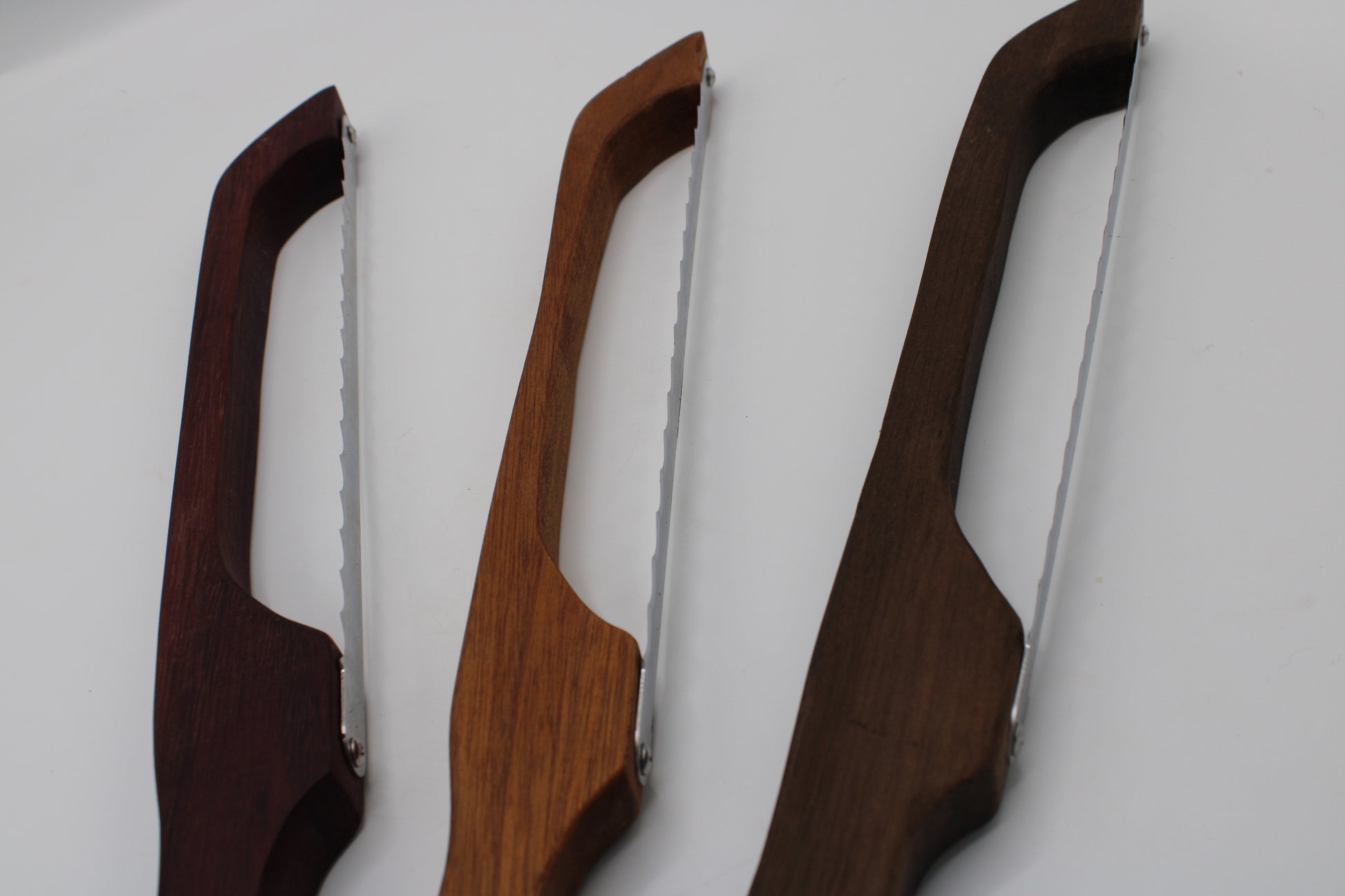 Three wooden-handled folding knives with serrated stainless steel blades, shown in three different wood finishes against a plain white background.