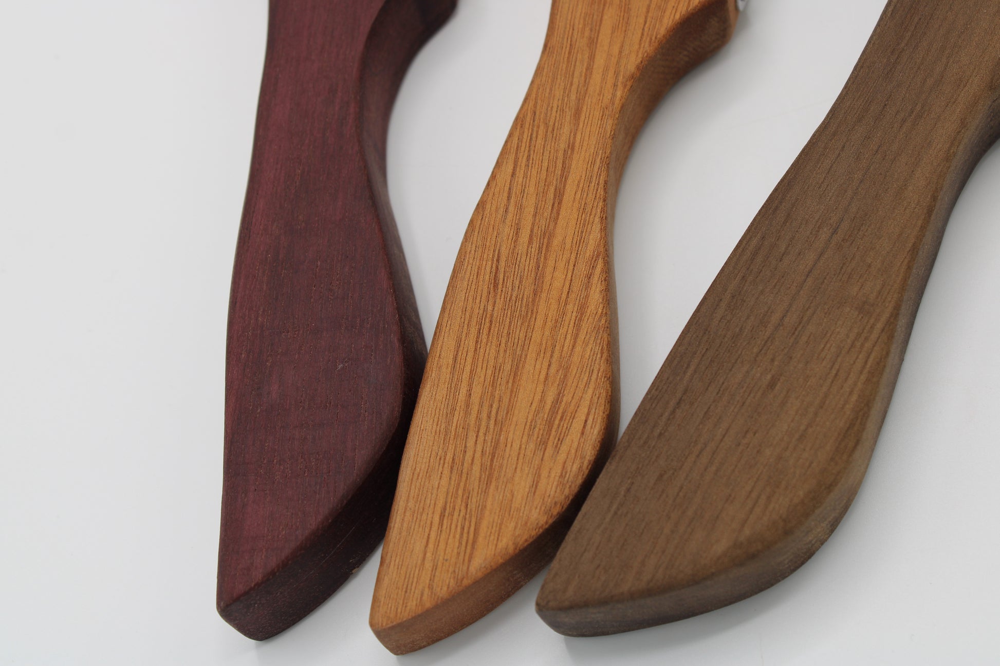 Three wooden serving utensils with curved handles and wide flat heads, shown in three different wood tones: dark burgundy, medium tan, and dark brown, on a light gray background.