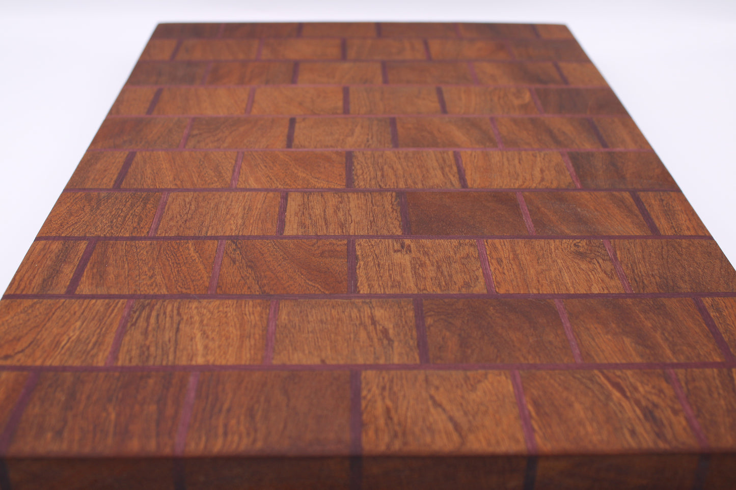 A rectangular wooden cutting board with a herringbone block pattern, featuring warm reddish-brown wood tones and dark purple end grain, shown on a white background.