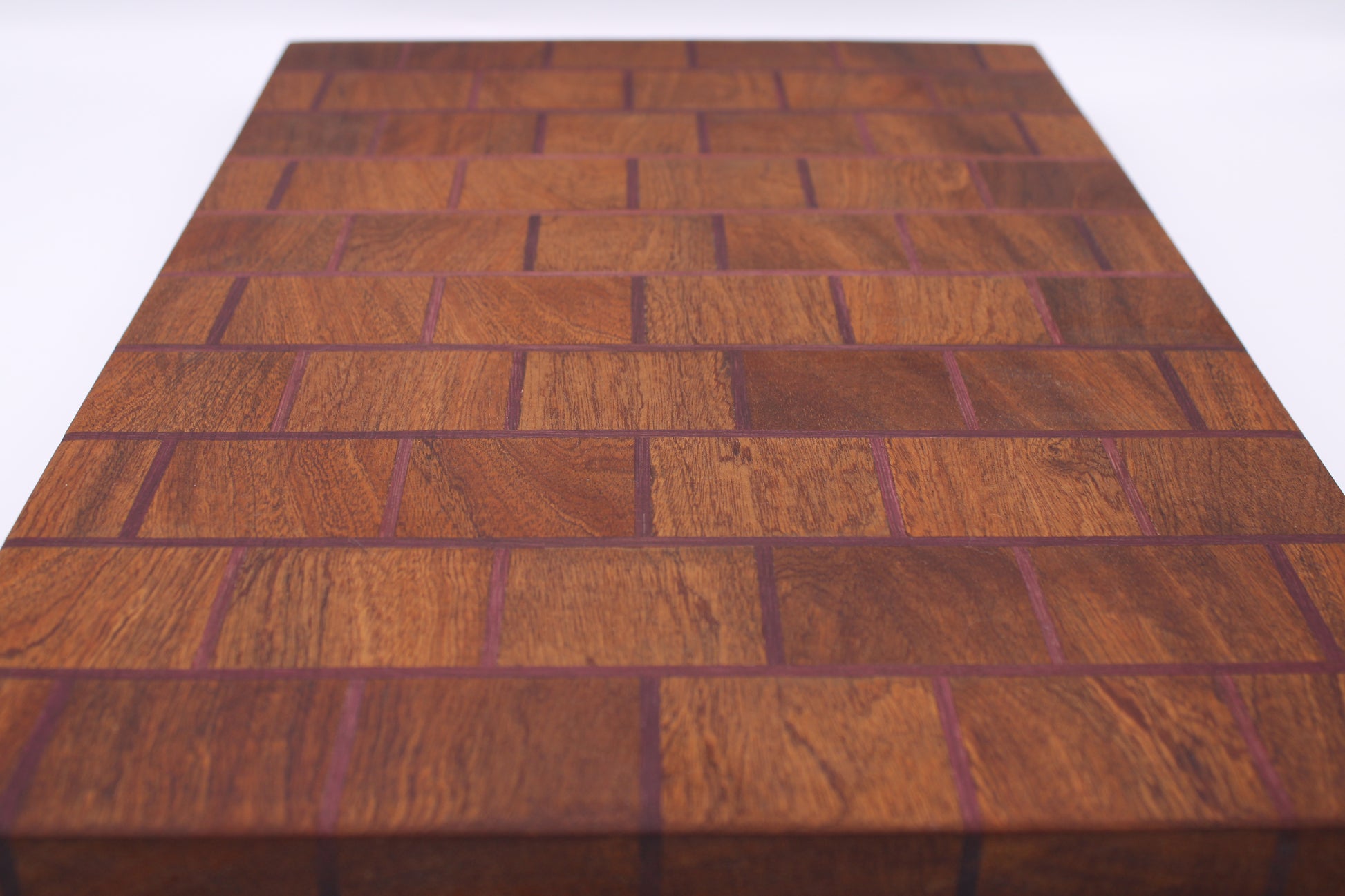 A rectangular wooden cutting board with a herringbone block pattern, featuring warm reddish-brown wood tones and dark purple end grain, shown on a white background.
