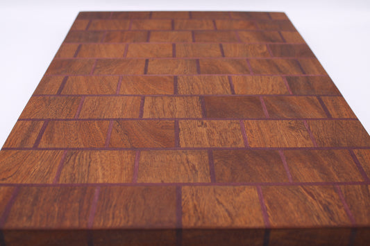 A rectangular wooden cutting board with a herringbone block pattern, featuring warm reddish-brown wood tones and dark purple end grain, shown on a white background.