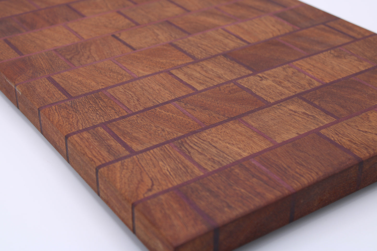 A close-up of a rectangular wooden cutting board made from interlocking wood blocks in a herringbone pattern, showing warm brown wood grain and dark reddish-brown seams, photographed on a white background.