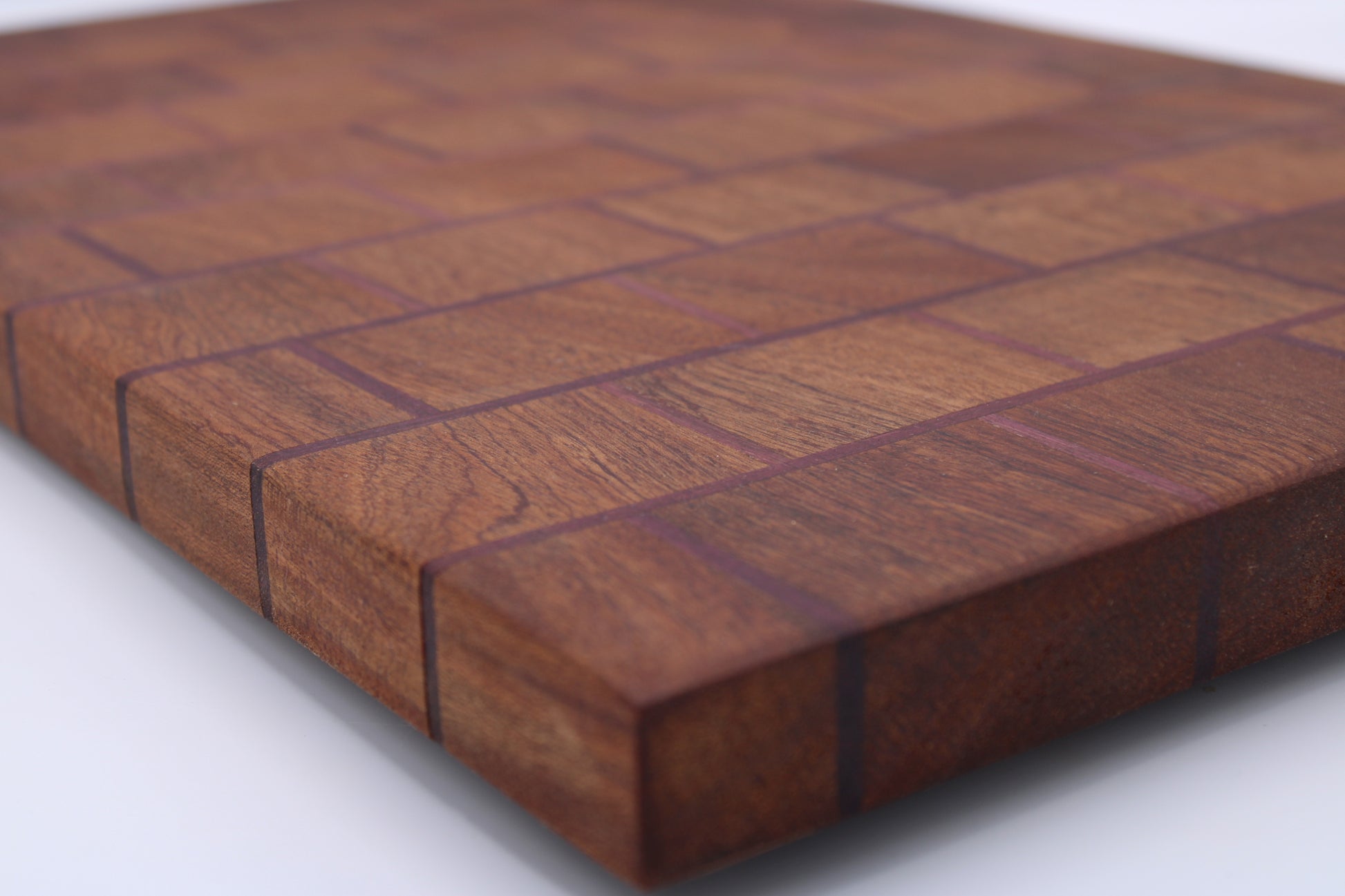 A close-up of a rectangular wooden cutting board made from interlocking wood blocks in a herringbone pattern, showing rich warm brown grain and dark dividing lines, photographed on a white surface.