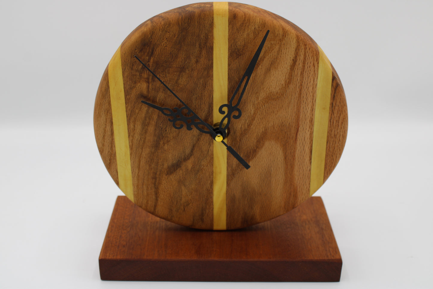 A round wooden clock with a natural wood grain face, accented by two vertical light wood stripes, black ornate hands, and a matching dark wood base on a white background.