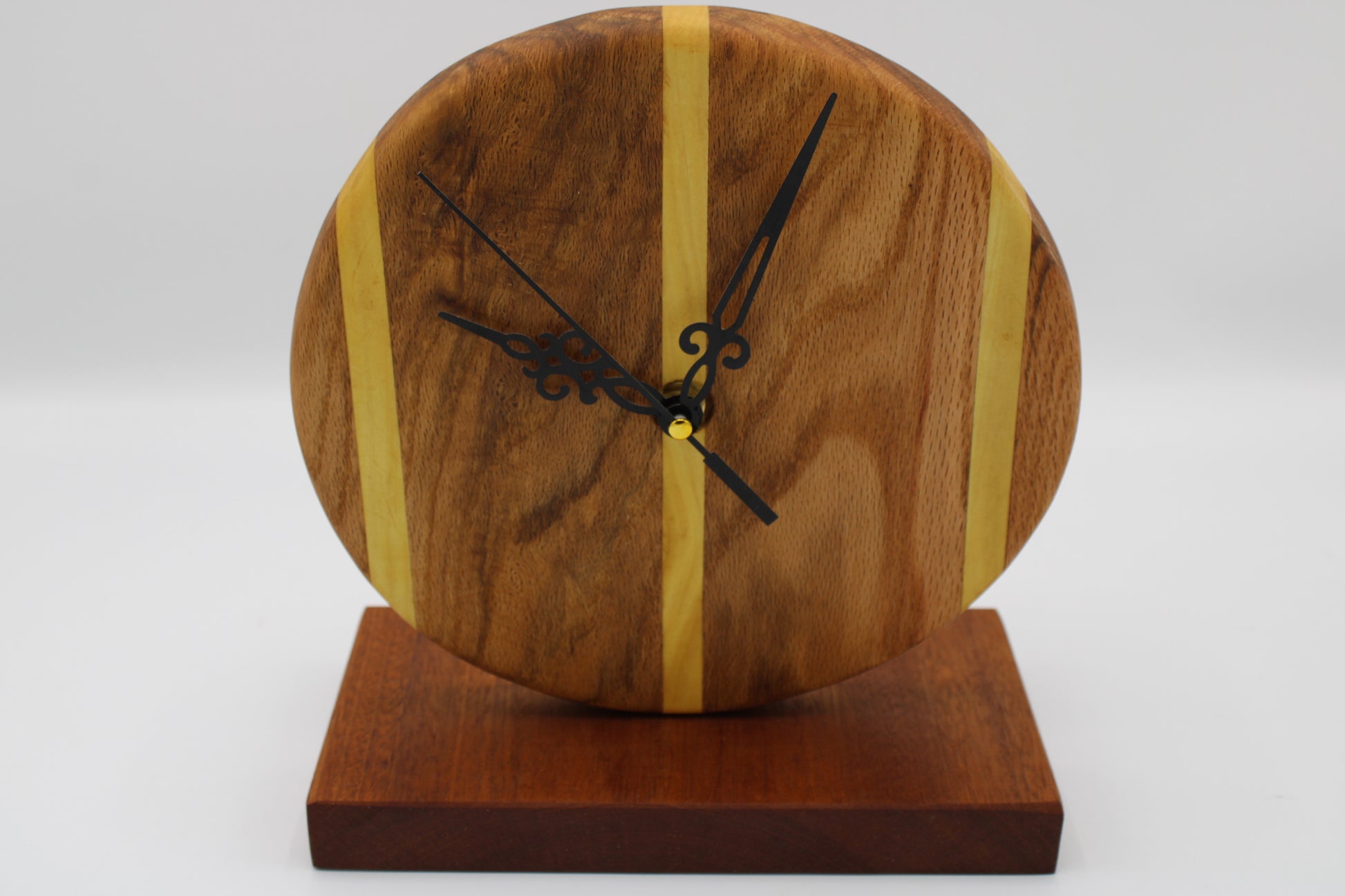 A round wooden clock with a natural wood grain face, accented by two vertical light wood stripes, black ornate hands, and a matching dark wood base on a white background.