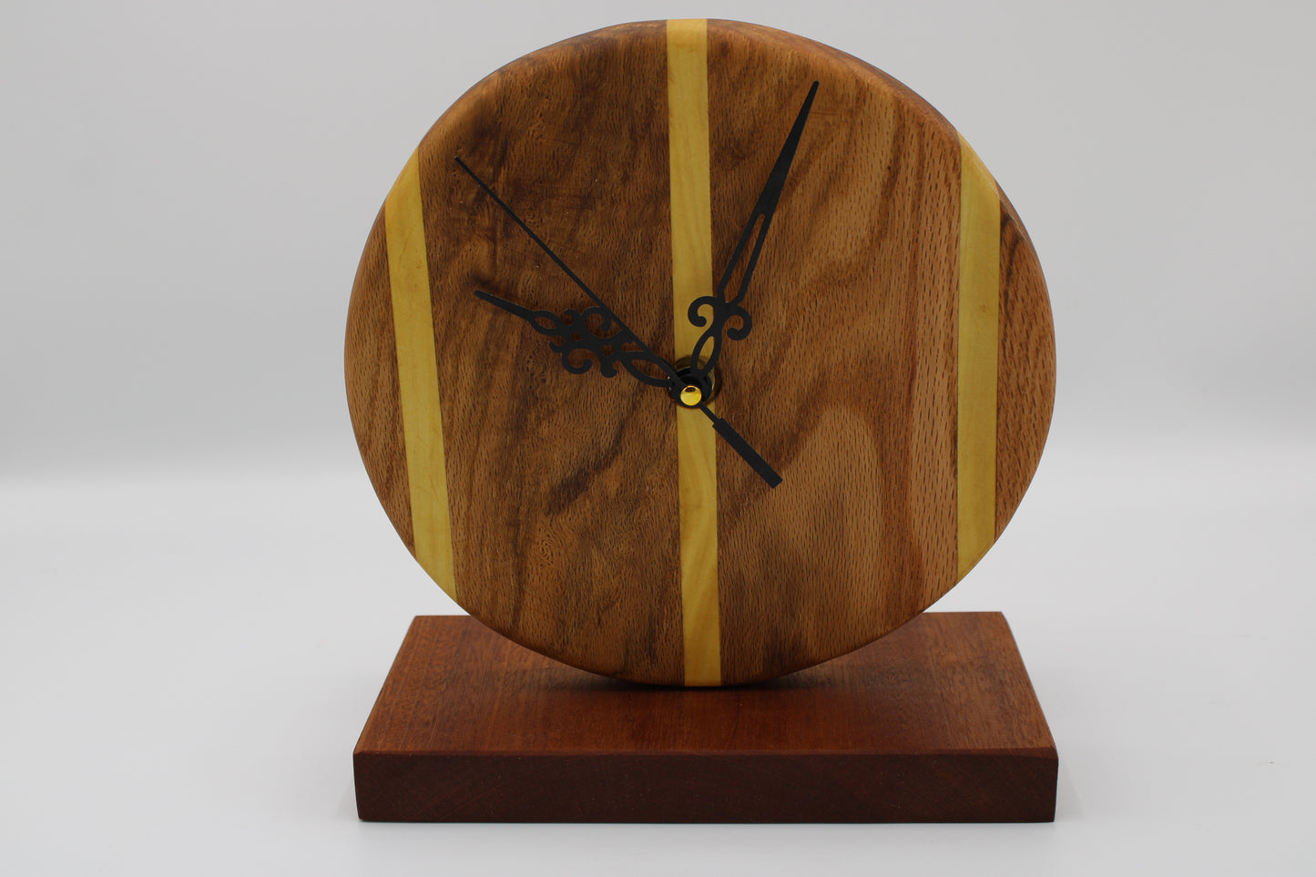 A round wooden wall clock with a natural wood grain face, accented by two vertical light wood stripes, black ornate hands, and a matching dark wood base on a light gray background.