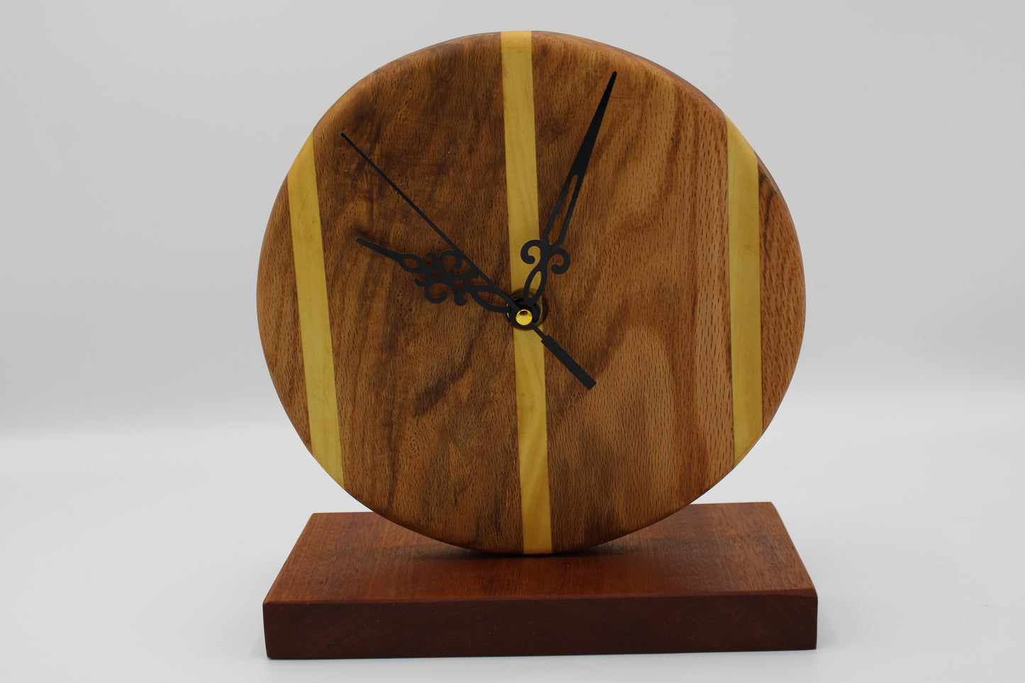 A round wooden wall clock with black ornate hands and vertical light wood inlays, mounted on a rectangular dark wood base against a light gray background.