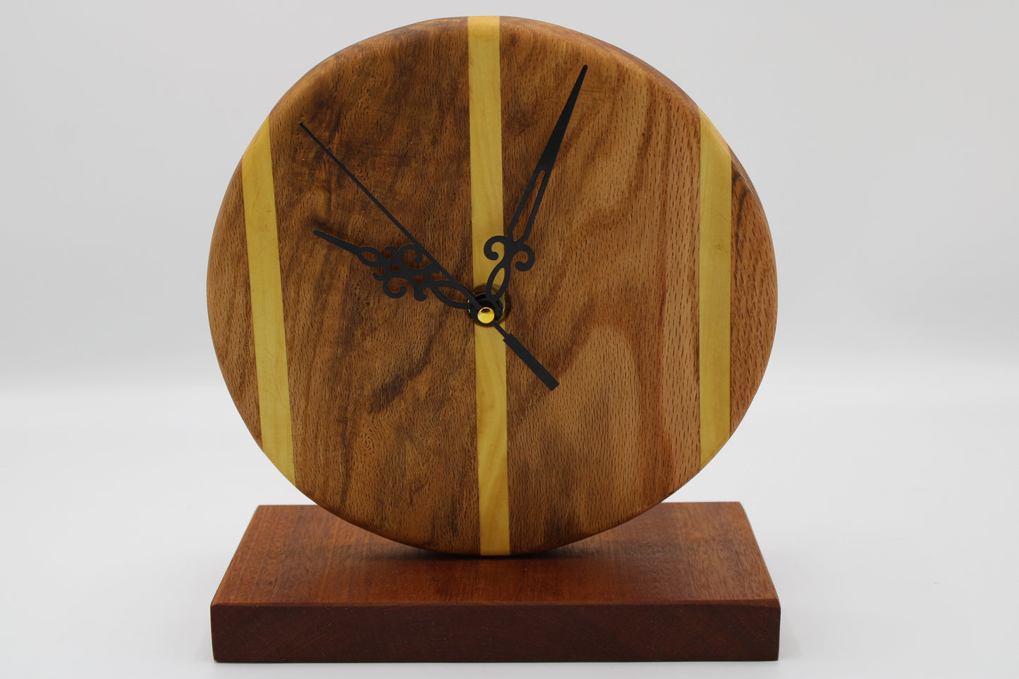 A round wooden wall clock with a natural wood face, two vertical light wood stripes, black ornate hands, and a dark brown wooden base, shown against a neutral light gray background.