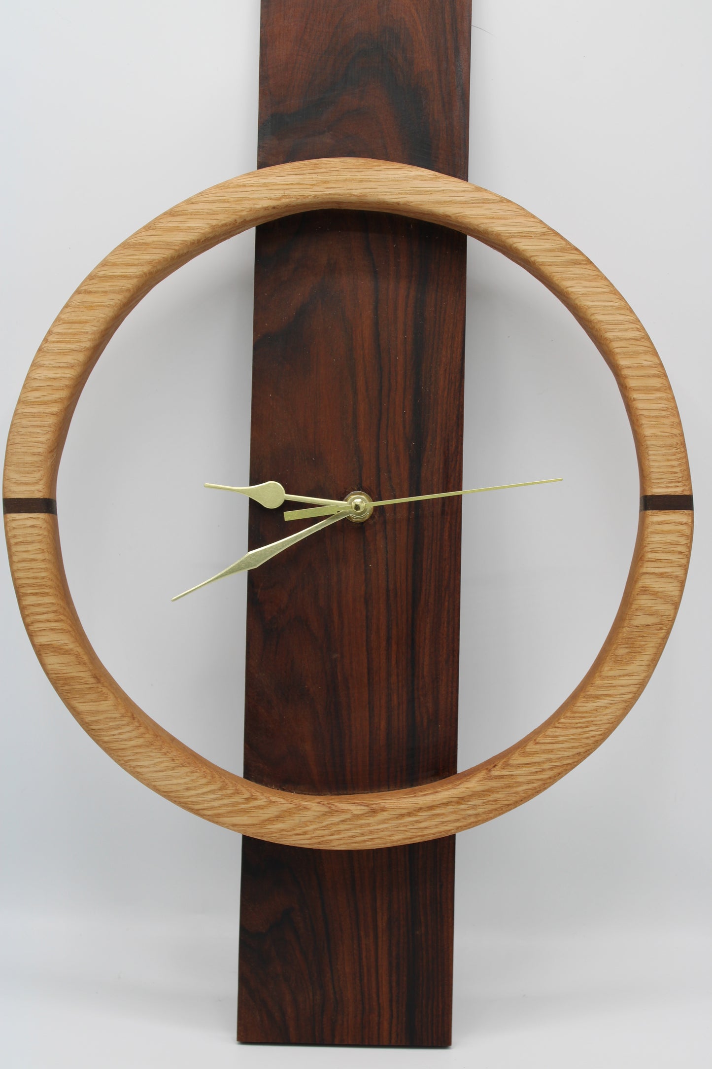A minimalist wall clock featuring a light oak circular frame and a dark brown wood-grain vertical rectangular base, with slim gold-toned hands.