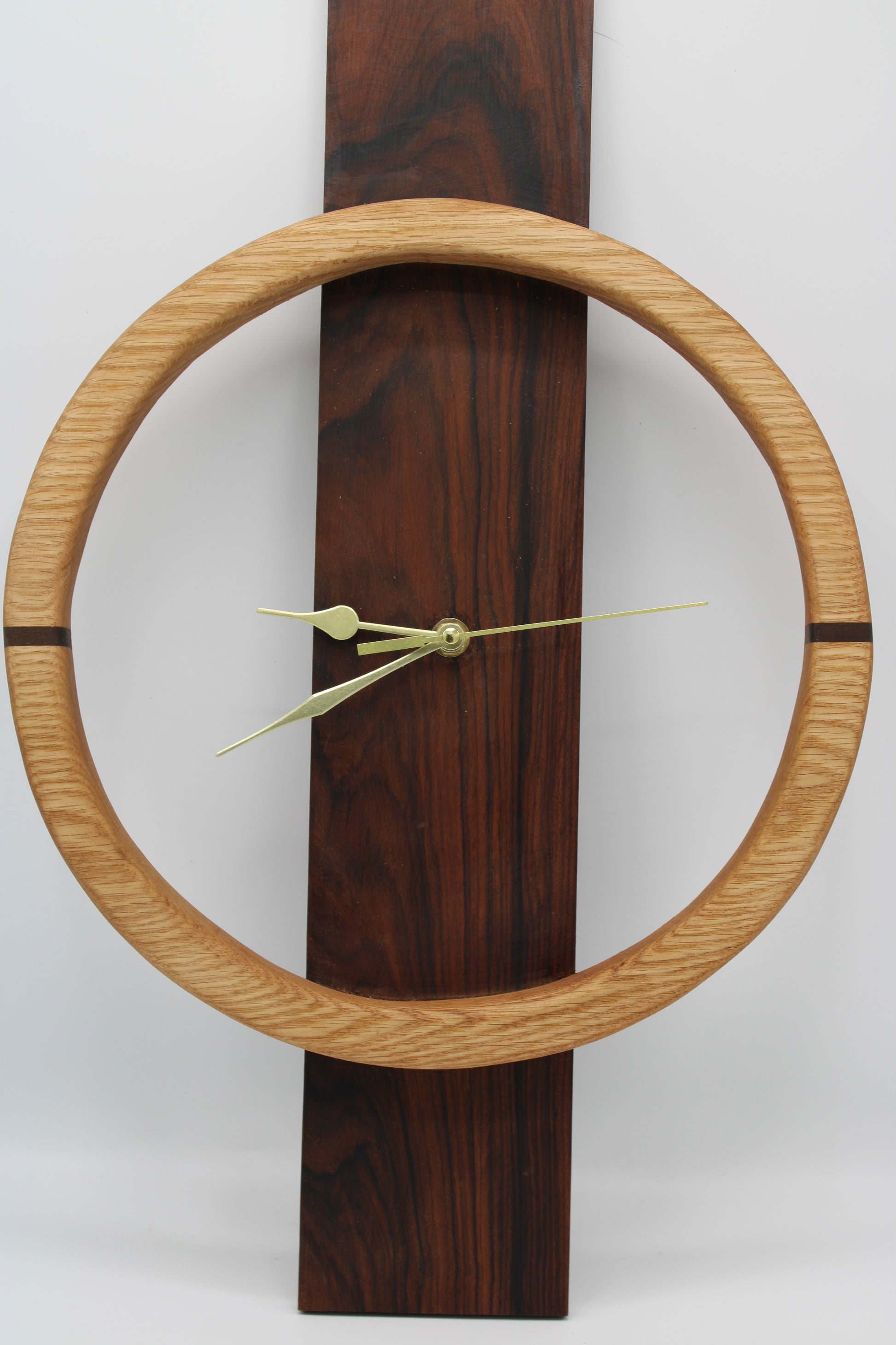 A minimalist wall clock featuring a light oak circular frame and a dark brown wood-grain vertical rectangular base, with slim gold-toned hands.