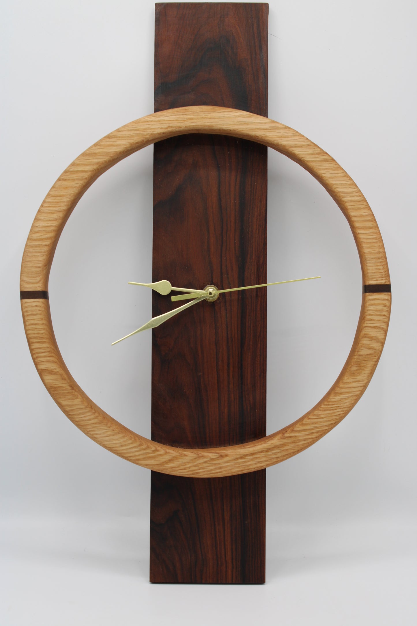 A minimalist wall clock featuring a large light oak circular frame around a dark reddish-brown wooden vertical plaque, with slim brass hands.