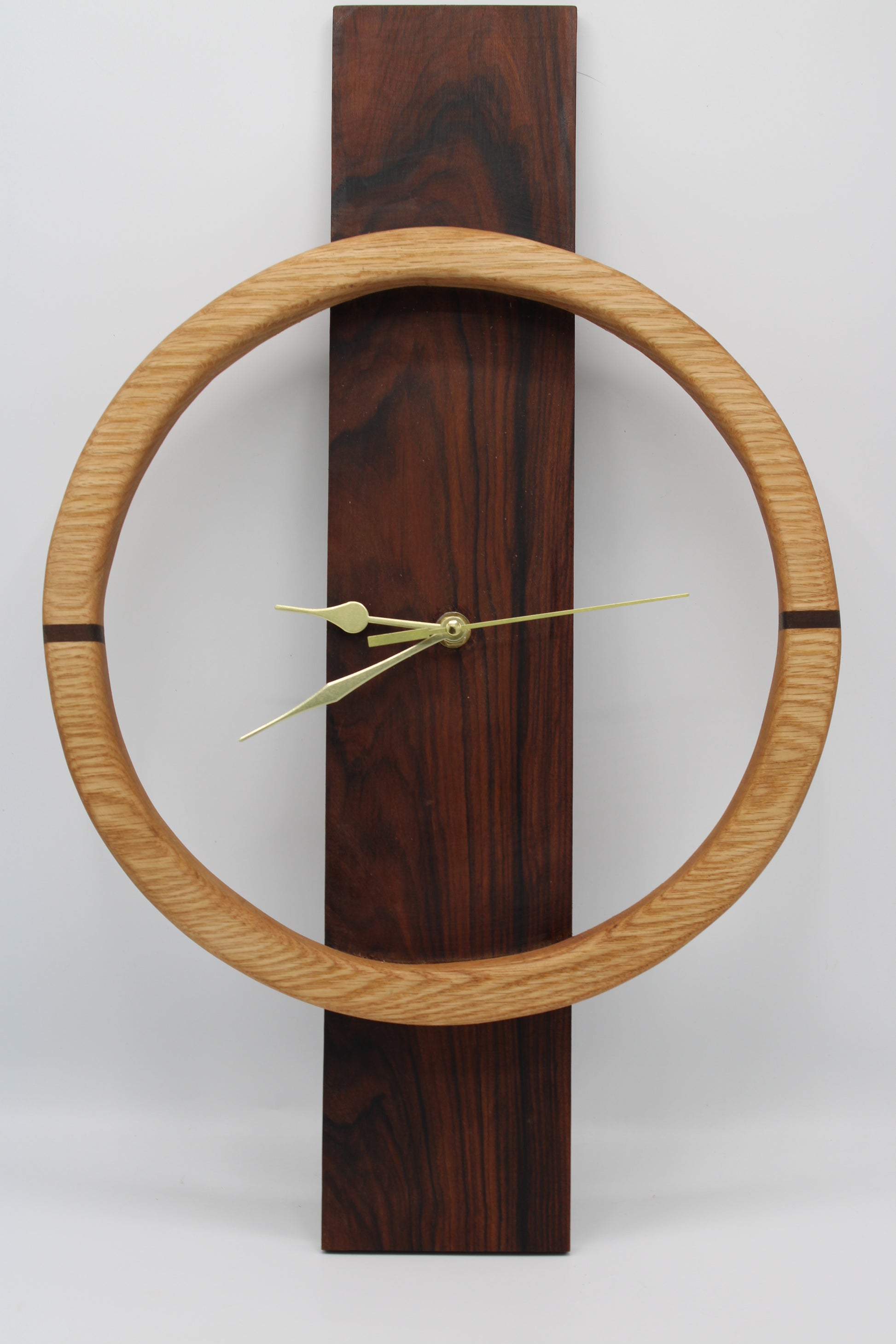 A minimalist wall clock featuring a large light oak circular frame around a dark reddish-brown wooden vertical plaque, with slim brass hands.
