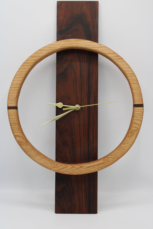 A minimalist wall clock featuring a large light oak circular frame around a dark reddish-brown wooden vertical plaque, with slim brass hands.