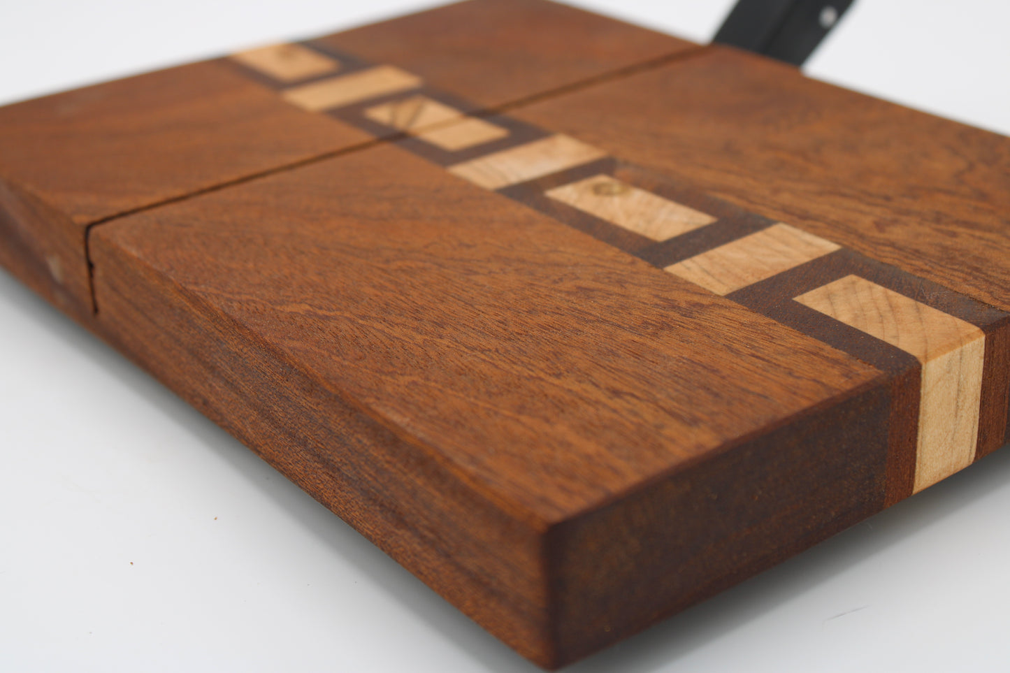 A close-up of a rectangular wooden cheese board with a rich dark brown wood grain and a geometric inlay pattern made of contrasting light tan wood strips, set against a plain white background