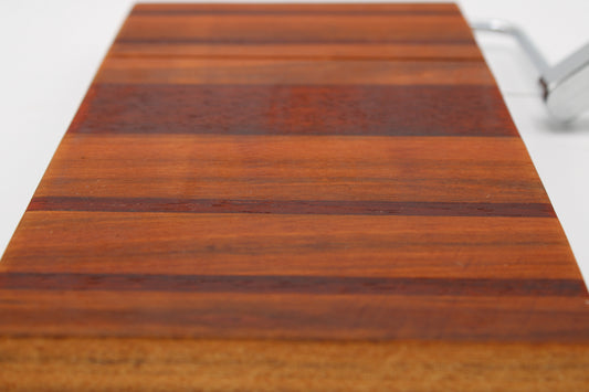 A close-up of a rectangular wooden cheese board with alternating medium and dark brown wood grain stripes, photographed on a white background with a small silver object partially visible at the top right.