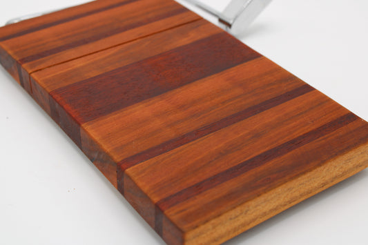 A close-up of a rectangular wooden cheese board with alternating dark and medium brown wood grain stripes, shown on a white background with a metal handle partially visible at the top.