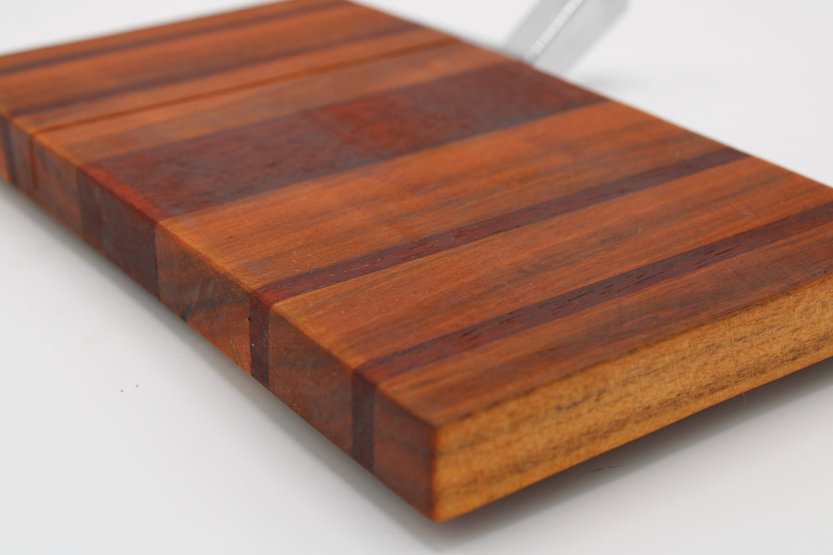 A close-up of a rectangular wooden cheese board with a striped grain pattern in warm brown and reddish tones, photographed on a white background.