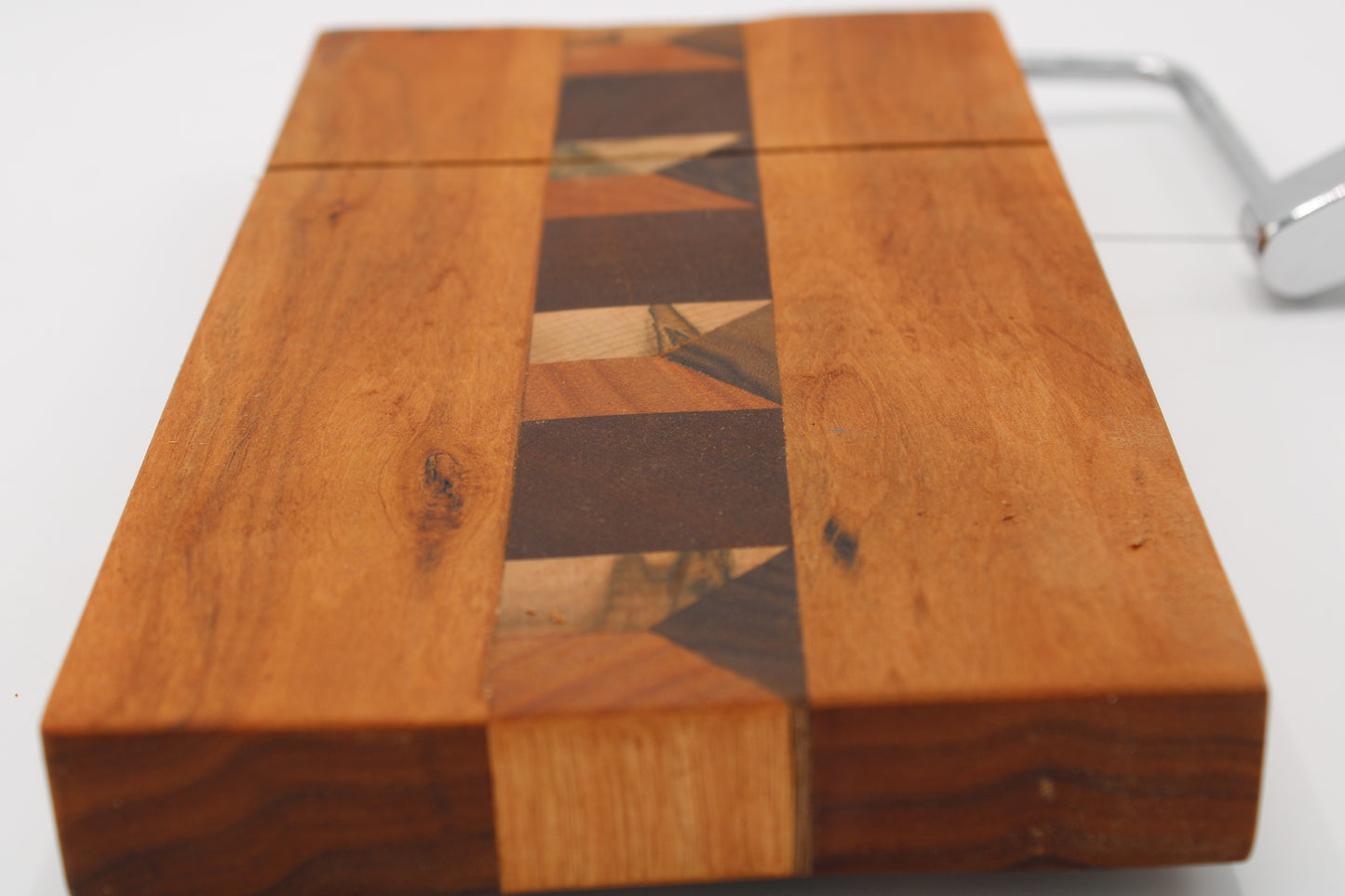 A close-up of a wooden cheese board with slicer with a geometric inlay pattern on the top, showing warm-toned wood panels and a silver metal handle on a white background.