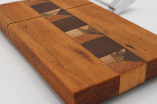 A close-up of a wooden cheese board with a geometric inlay pattern made of dark and light wood pieces, photographed on a white background.