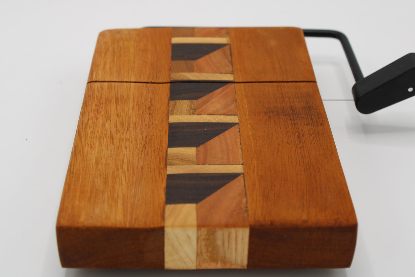 A square wooden cheese board with a geometric inlay pattern down the center, made from multiple shades of natural wood, shown on a white background with a black handle visible on the right.
