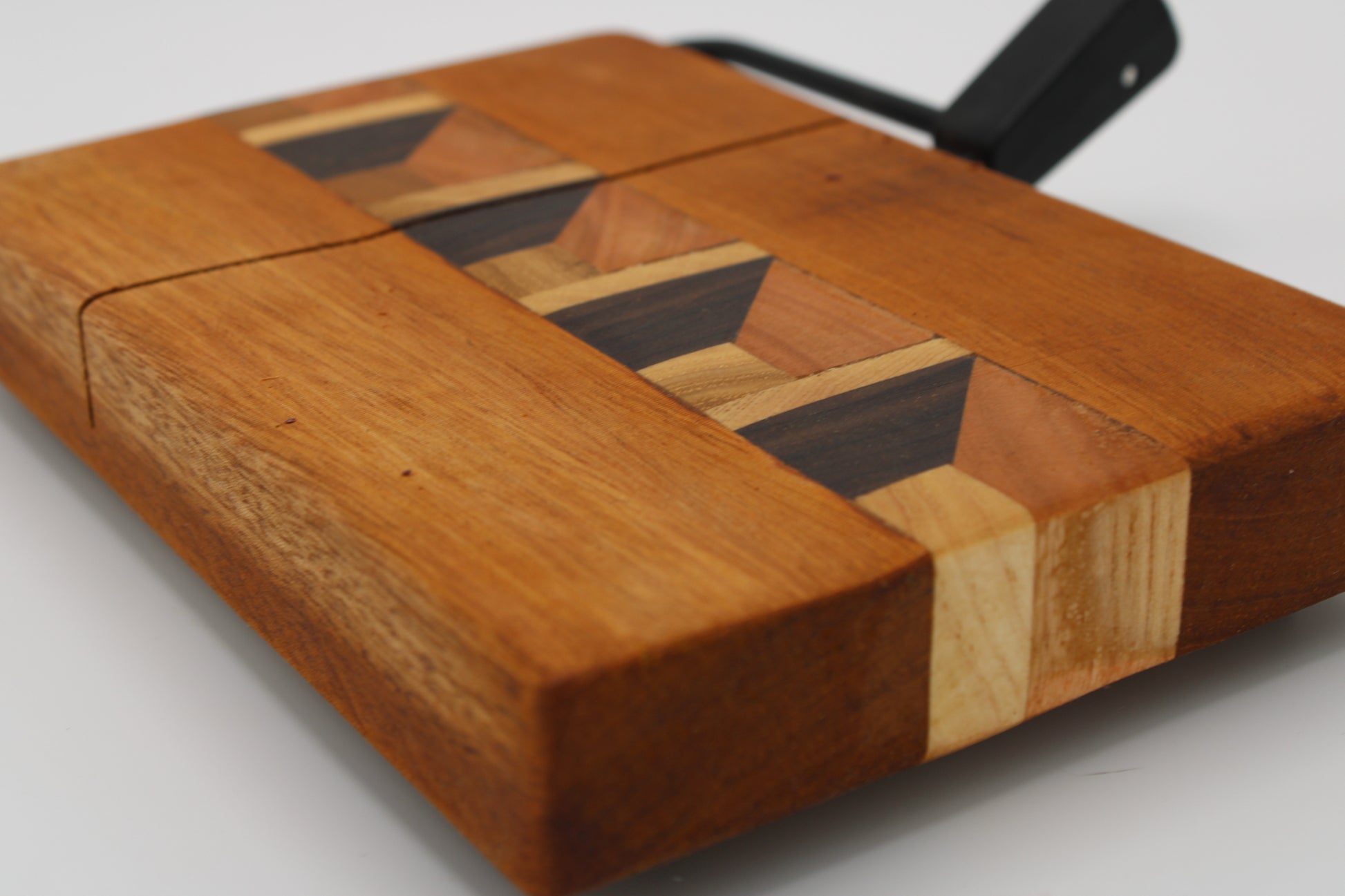 A close-up of a wooden cheese board with a geometric inlay pattern made of light and dark wood tones, featuring a black metal handle on a white background.