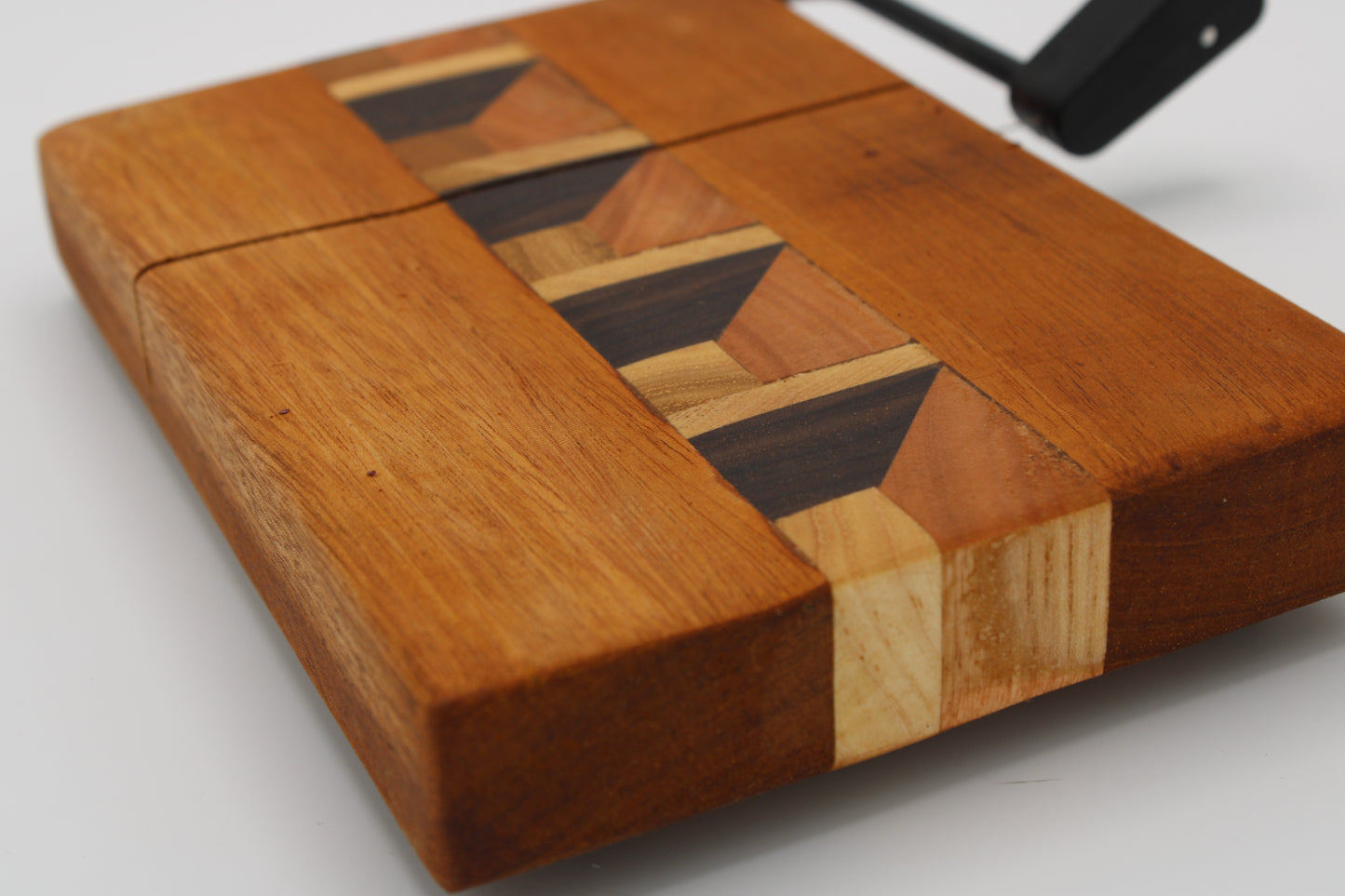 A wooden cheese board with a geometric inlay design, featuring a warm brown wood body and a central pattern of dark and light wood strips, shown on a light gray background.